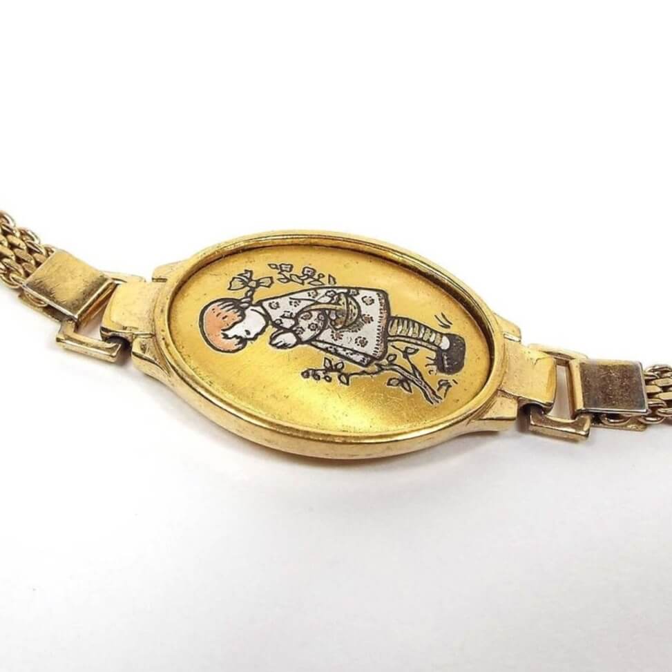 Top view of the retro vintage Reed and Barton Damascene bracelet. The middle of the bracelet has a large oval with a scene of a girl with her head bent and hands together praying outside. Behind her is a branch. Her hair is copper color and her dress is painted white. The rest of the bracelet is gold tone in color. At the top and bottom of the oval part is a rectagle ring for the rest of the bracelet to attach to. The side straps are mesh chain design with a snap lock clasp at the end.
