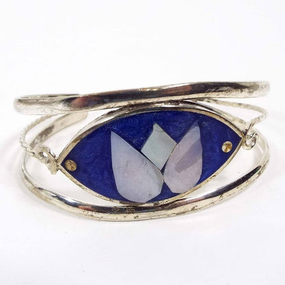Front side of the Alpaca Mexico cuff bracelet has a curved tapered oval with bright pearly blue resin enamel. In the middle is a large lotus flower design with pearly mother of pearl shell petals. There is a curved band of darkened silver color metal on the top and bottom and a thinner twisted and curled band of metal in the middle coming off the sides of the oval to form the sides and back of the cuff. 