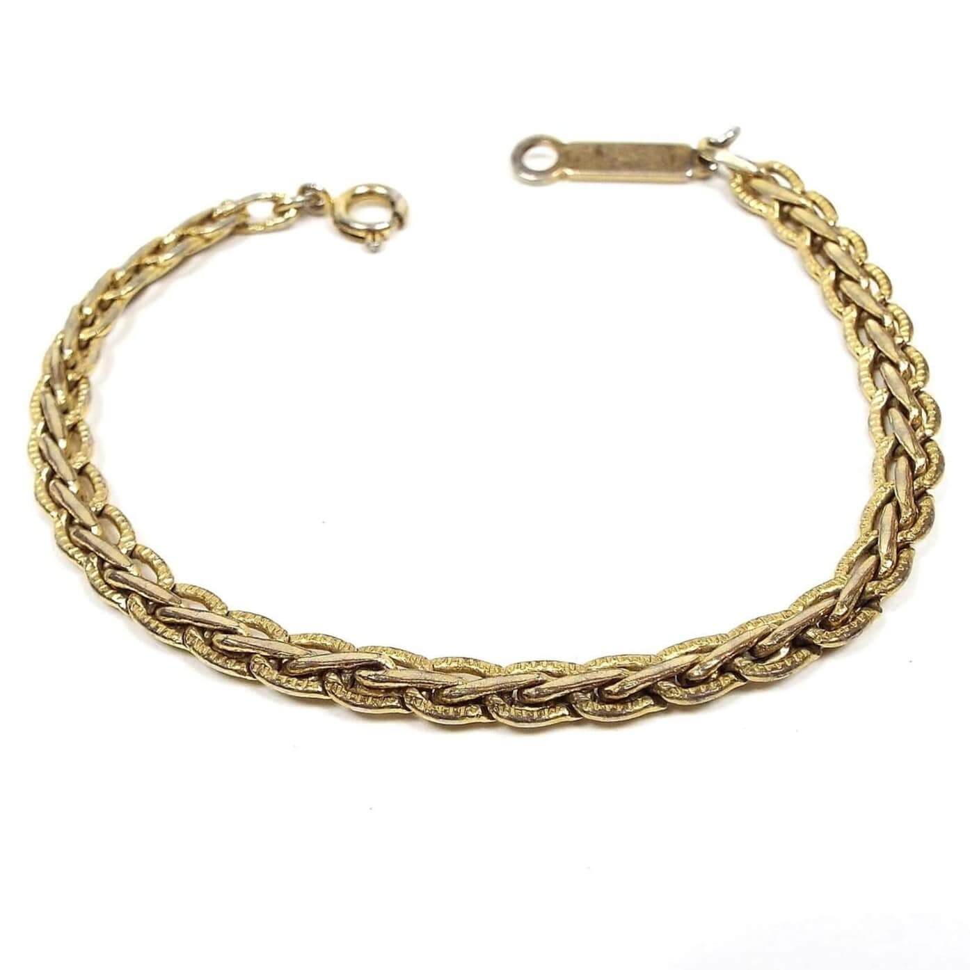 Angled top view of the retro vintage fancy link chain bracelet. It is a slightly darkened gold tone in color. There is a spring ring clasp on one end and a long bar with a loop on the other. The link design is a wheat style braid.