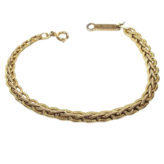 Angled top view of the retro vintage fancy link chain bracelet. It is a slightly darkened gold tone in color. There is a spring ring clasp on one end and a long bar with a loop on the other. The link design is a wheat style braid.