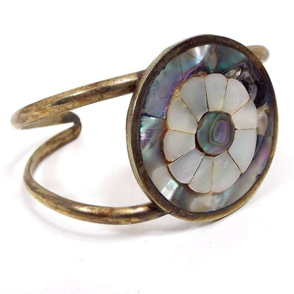 Front and side view of the Alpaca marked cuff bracelet. Bracelet has a large round middle piece with flower design. Flower petals are pearly white mother of pearl shell and the rest of the circle is pearly multi color abalone shell. Two rounded bands of metal come off each side and curve to the back to form the cuff. Metal is a darkened silver in color. 