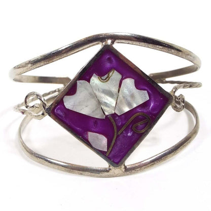 Front view of the Mexican alpaca floral cuff bracelet. There are three curved bands of metal forming the cuff part of the bracelet. The top and bottom have rounded fronts. The middle wire is twisted and curls at the ends by the focal point of the bracelet. The middle has a large diamond shape with pearly purple resin enamel. There is a pearly white lotus flower design with inlaid mother of pearl shell for the leaf and petals. The metal is silver tone in color.