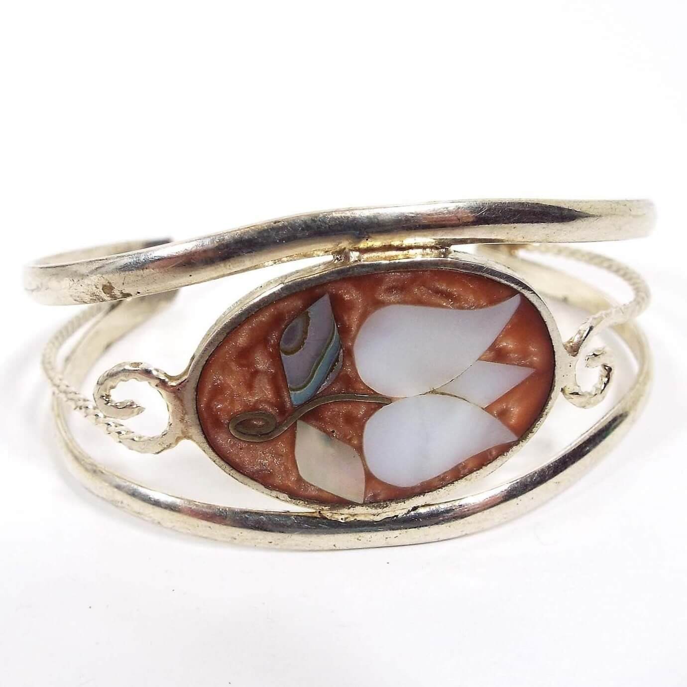 Picture of the front of the Alpaca cuff bracelet. Middle of the bracelet has a large oval with dark pearly peach color resin. Inside the resin is a flower design with mother of pearl petals and abalone shell leaves.It makes the flower petals look white and the rest of the flower multi color. The oval has a band of silver color metal above and below it forming the curved cuff of the bracelet. On each side of the oval is a thinner twisted band of silver color metal that is curved around the sides.