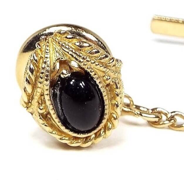 Front view of the Mid Century vintage Swank tie tack. The metal is gold tone in color. There is a small oval black glass cab in the middle. The bottom has a twisted rope style design curved around the bottom of the cab. The top has another twisted rope style design that is angled like a V shape over the top of the cab area. The round clutch on the back has a chain with a small bar on the end.