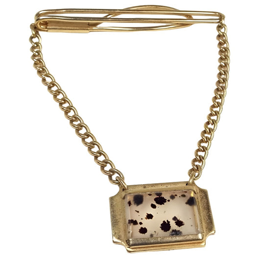 Front view of the 1940's Mid Century vintage gemstone tie bar chain. The metal is gold tone in color. The front bar is a long open oval and the back bar has a bar and then a wider open oval area for a slide on style tie bar. There is curved oval curb chain coming down from either side with a wide octagon shaped charm at the bottom. The inside of the charm has a rectangular dendric agate slab that is clear with black splotchy spots running throughout it.