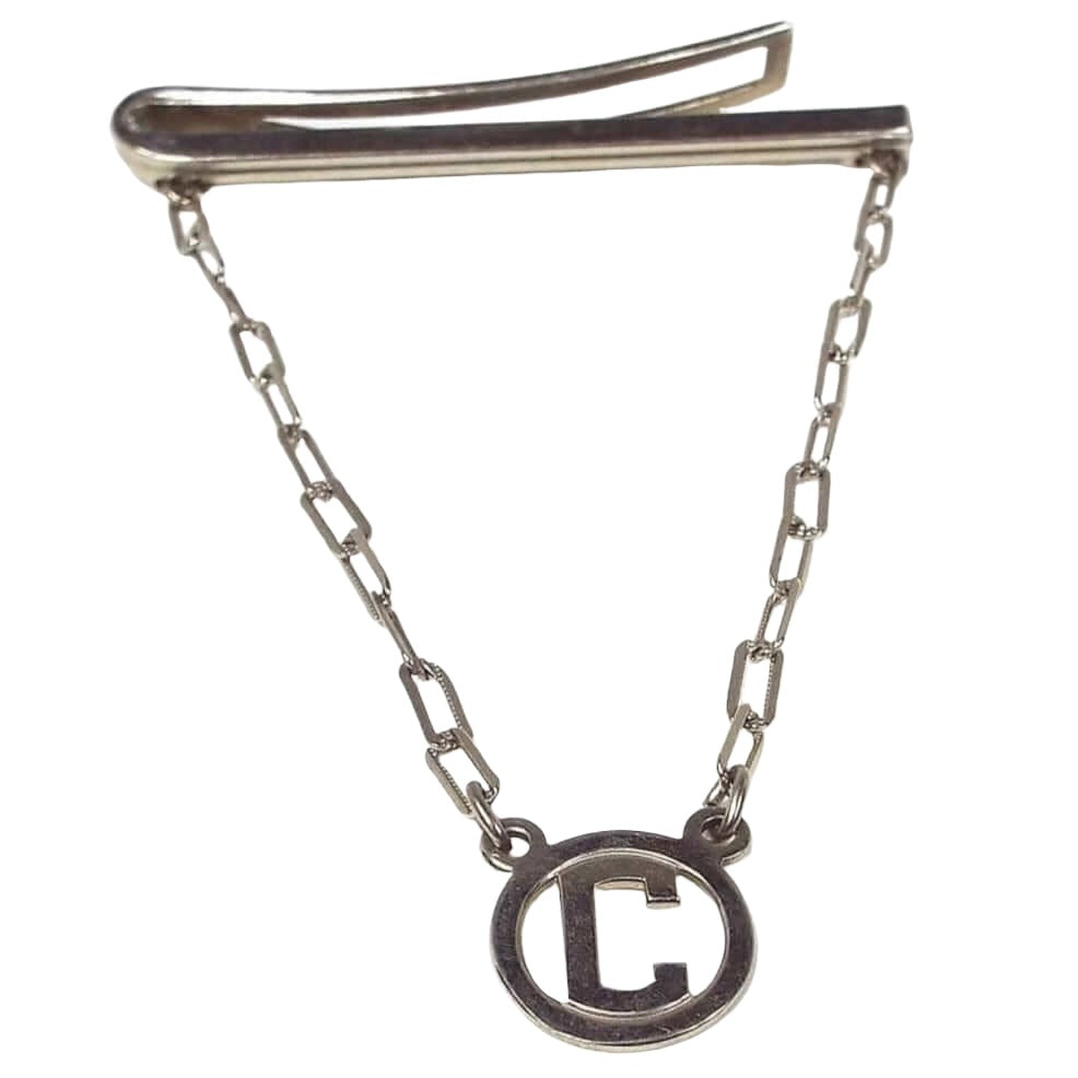 Angled front and side view of the Mid Century vintage tie bar chain. The metal is silver tone in color. The side view of the top bar shows a line design on the front and a curved back bar with open middle. The back bar is cuved outwards some, so it would be better for thicker style ties. Small cable chain with rectangle links comes down from each side of the bar and there is a round open charm at the bottom with a block letter initial C in the middle.