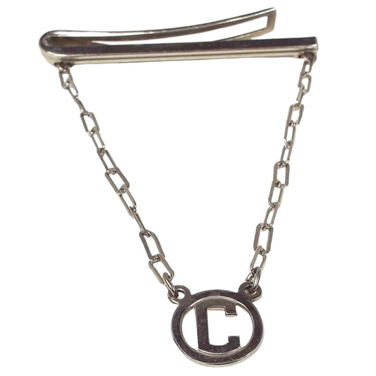 Angled front and side view of the Mid Century vintage tie bar chain. The metal is silver tone in color. The side view of the top bar shows a line design on the front and a curved back bar with open middle. The back bar is cuved outwards some, so it would be better for thicker style ties. Small cable chain with rectangle links comes down from each side of the bar and there is a round open charm at the bottom with a block letter initial C in the middle.
