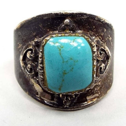 Top view of the retro vintage CFJ sterling silver turquoise ring. The sterling is heavily darkened from age for a dark gray color. It is wide on the top and tapers down the sides. The middle has a puffy rounded square turquoise gemstone cab that is blue in color with tiny thin light brown lines running through it. There are some small fine fissures when viewed under magnification. There is a spade like design on each side of the stone.  