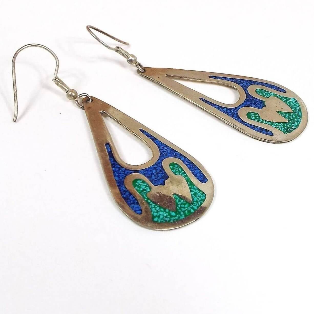 Front view of the Alpaca earrings are teardrop shaped with smaller tear drop cut outs at the top part of the earrings. There are two wavy areas of resin at the bottom. Each part has tiny stone chips inlaid in the resin. The top part is blue and the bottom part is green. The metal on the earrings is silver in color and they have curved wire fish hook style ear wires at the top.