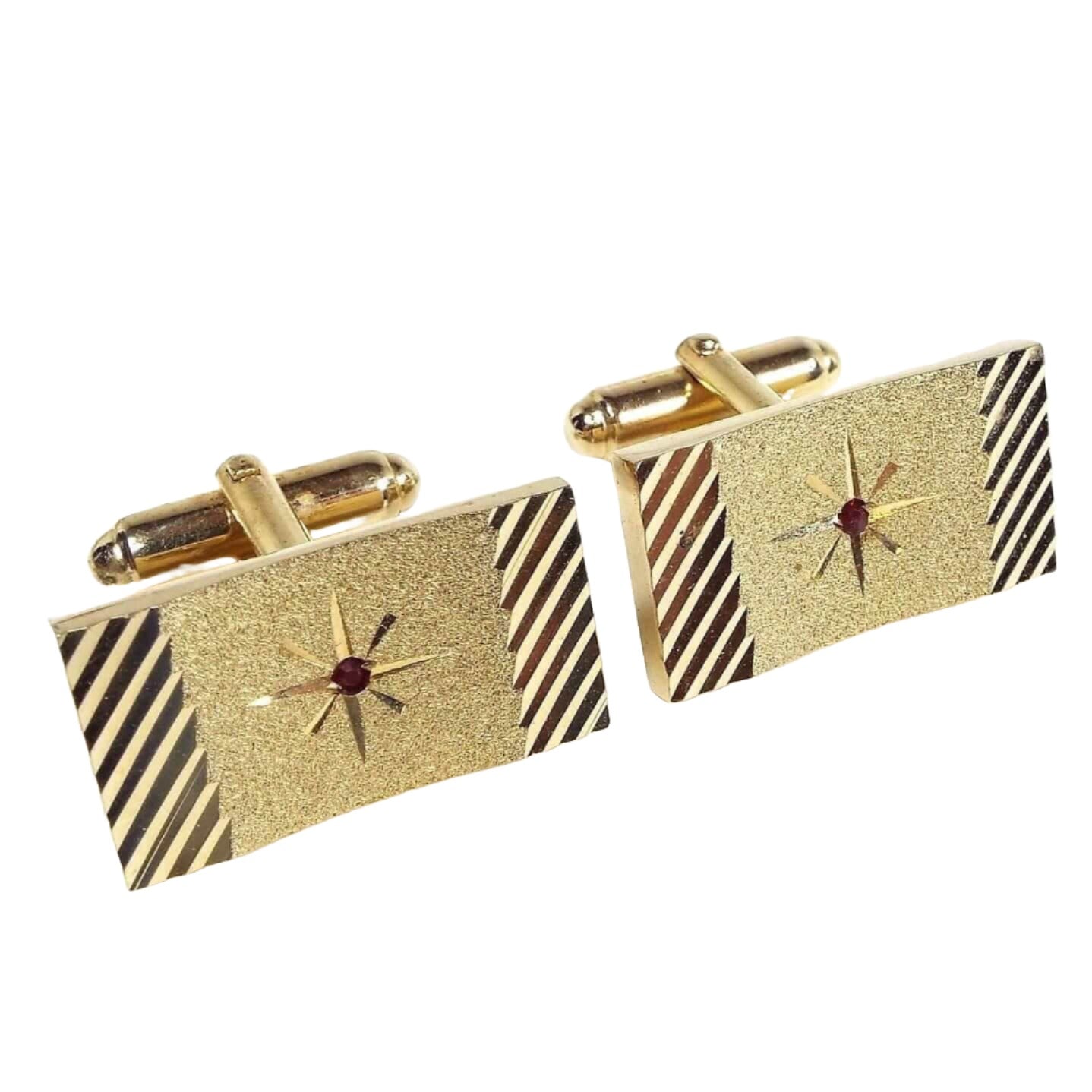 Angled front view of the Mid Century vintage Anson cufflinks. They are gold tone in color and rectangle in shape. The fronts have a cut etched starburst design with a single red rhinestone in the very middle. The sides have a diagonal cut etched line design. 