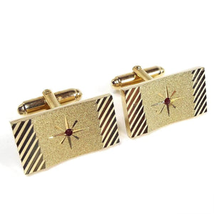 Angled front view of the Mid Century vintage Anson cufflinks. They are gold tone in color and rectangle in shape. The fronts have a cut etched starburst design with a single red rhinestone in the very middle. The sides have a diagonal cut etched line design. 