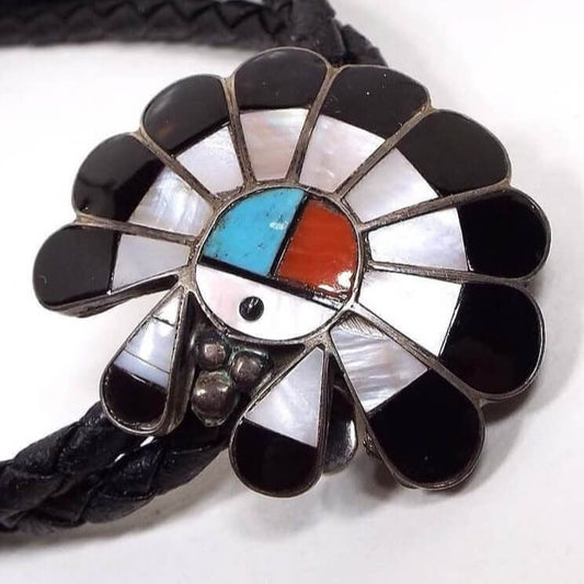 Front view of the retro vintage Bennett Zuni Sun Face bolo tie slide. Each feather in the Chief headdress design has inlaid pearly white mother of pearl with inlaid black onyx gemstone at the ends. The face area is a circle that has inlaid mother of pearl on the bottom and a small dot of onyx for the mouth. The top part has inlaid blue turquoise on the left and the right side has inlaid red color coral. Cord is braided dyed black leather with darkened silver tone tapered ends.