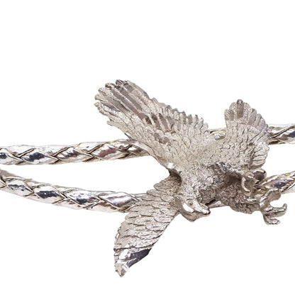 Zoomed in front view of the retro vintage eagle bolo tie. The slide depicts a detailed flying eagle with its talons out and is silver tone in color. The imitation leather braided cord is metallic silver color. There are two silver color metal cone ends at the end of the bolo tie.