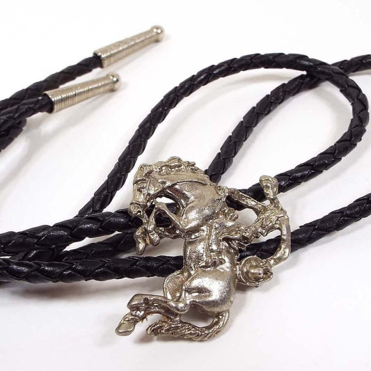 Front view of the retro vintage bolo tie. The bolo slide is made of pewter and is silvery gray in color. It has a semi 3D appearance depicting a cowboy riding a bucking bronco. The man is holding his hat in one hand and the reins in the other. The bolo tie itself has black woven cord with tapered silver tone cone ends.