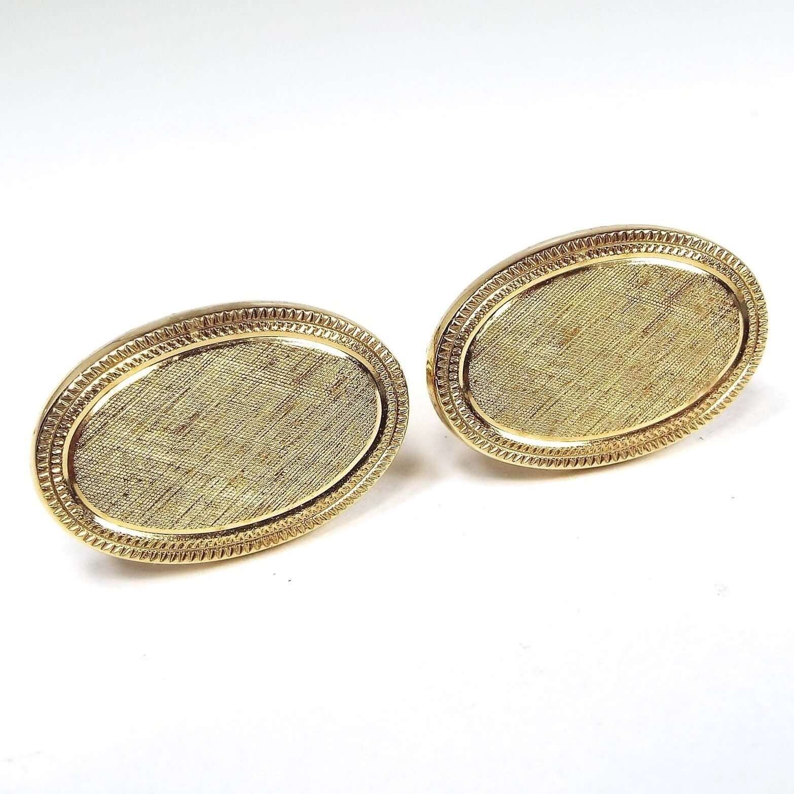 Front view of the retro 1970's Avon cufflinks.The metal is gold tone in color. They are oval in shape with brushed textured matte middle areas. The outer edge has has a slightly raised shiny gold tone edge and two rows of etched lines. 