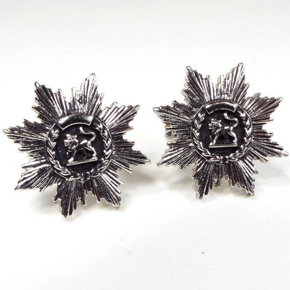Front view of the Mid Century vintage Swank lion cufflinks. the metal is antiqued silver tone in color. There is a round area in the middle with a side view of a lion on a black painted background. There is a wheat style design curved around the bottom edge. All the way around the cufflinks is a brutalist starburst like area of line textured leaf like shapes. 