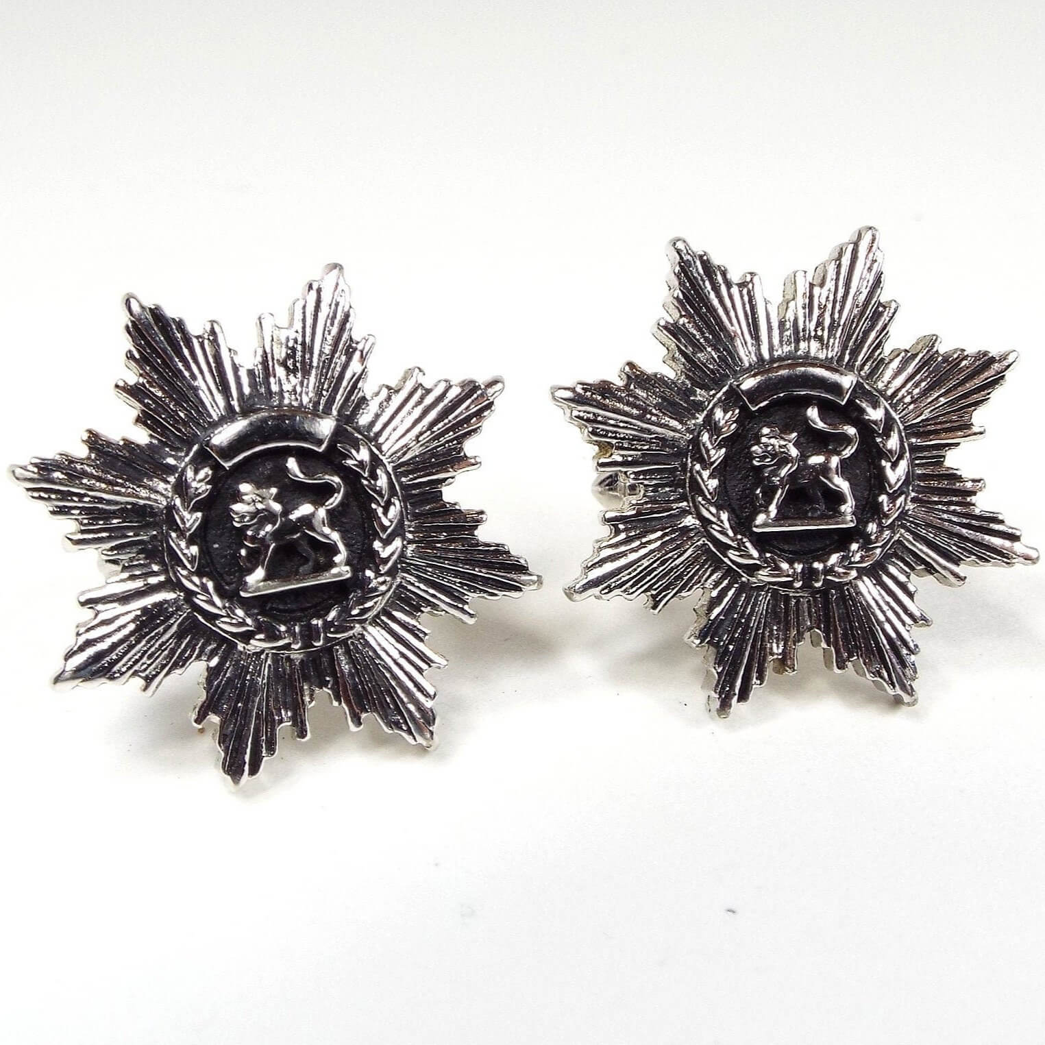 Front view of the Mid Century vintage Swank lion cufflinks. the metal is antiqued silver tone in color. There is a round area in the middle with a side view of a lion on a black painted background. There is a wheat style design curved around the bottom edge. All the way around the cufflinks is a brutalist starburst like area of line textured leaf like shapes. 
