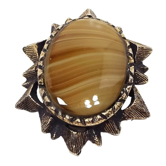 Brooch has a line textured metal in antiqued brass color edge with triangles coming off of it for a sun or star like design. In the middle is an oval agate gemstone cab that has thin wavy lines of yellow and brown color. The edge of the gemstone cab is faceted.