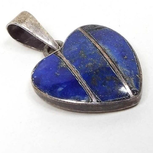 Angled front view of the small retro vintage gemstone heart pendant. The metal is silver tone in color. The pendant is heart shaped and has two diagonal lines of metal running through it. There are three pieces of inlaid lapis lazuli in between the metal lines and outer bezel area. The lapis lazuli is deep denim blue in color with tiny metallic flecks. There is a loop bail attached to the built in ring at the top of the pendant.