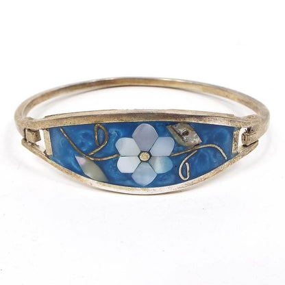 Front view of the Mexican Alpaca hinged bangle. It's silver in color with a curved metal band on the back and split double bands of metal on the front. In between is a curved shaped piece that has pearly blue resin. In the middle is a flower design with leaves and stem. The flower petals are mother of pearl shell and are pearly white color. The leaves are abalone shell and are pearly multi color. The stem is metal. One side has a metal hook style piece so you can open and close the bangle.  