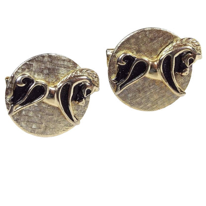 Front view of the Mid Century vintage Cathe cufflinks.They are round with brushed matte metal gold tone color. There are raised horse designs that look similar to a rocking horse without the bottom rocker part. They are gold tone and black painted in color.