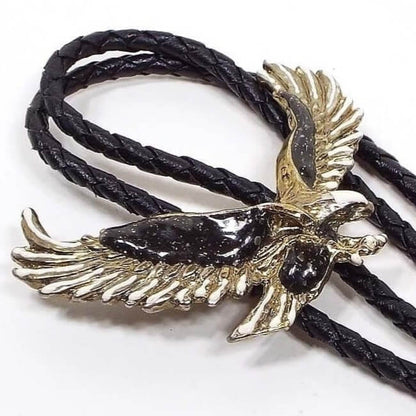 Front view of the retro vintage eagle bolo tie. The bolo slide is a textured flying eagle design. The eagle's body and wings have black enamel with confetti glitter embedded in it. The tips of the eagle's wings are enameled white. The metal is pewter and a light gray color. It has a black braided imitation leather cord. 