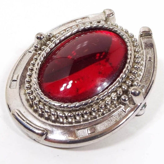 Front view of the retro vintage bolo slide. The middle has a large oval red glass cab that is bezel set. The bezel area has a twisted rope style design. Around that is a row of round dots. The outer area of the slide is shaped like a horseshoe. The metal is silver tone in color. This is just the slide and there is no cord bolo tie part with it.
