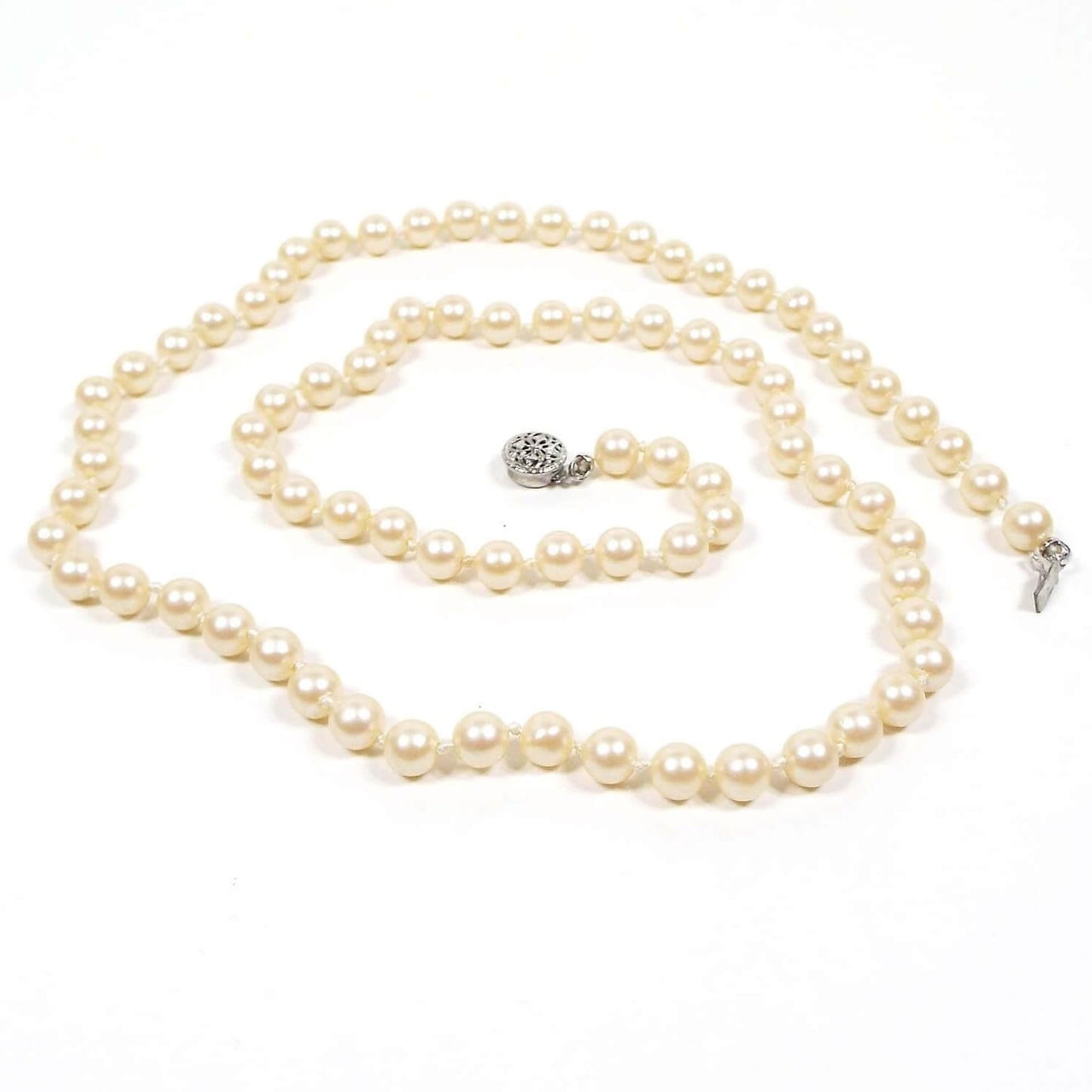 Top view of the Mid Century vintage faux pearl necklace. It is one strand of coated plastic round beads in an off white golden yellow color. The beads are have knots in between them. There is a silver tone filigree box clasp at the end.