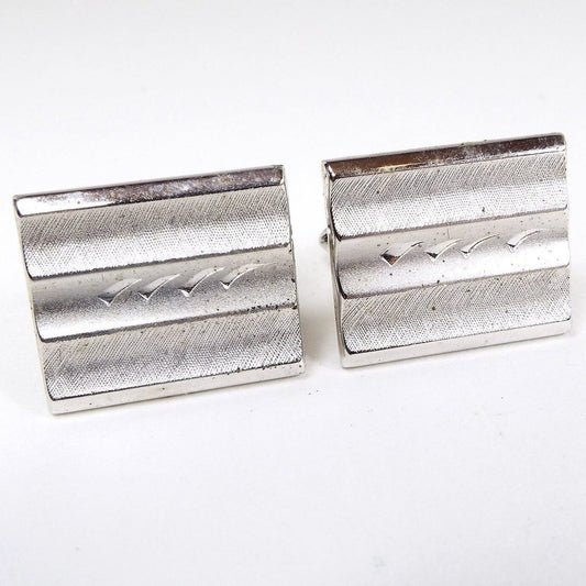Front view of the Mid Century vintage Swank Cufflinks. They are matte textured brushed silver tone in color and have a rectangle shape. The fronts have three rounded indented areas lengthwise and the middle one has an etched design of curved flares. 