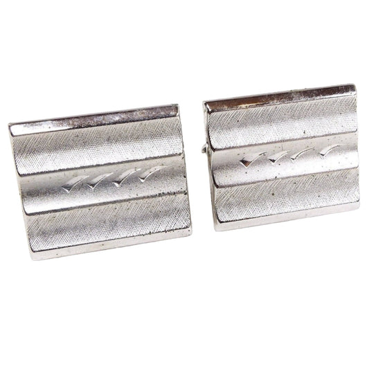 Front view of the Mid Century vintage Swank Cufflinks. They are matte textured brushed silver tone in color and have a rectangle shape. The fronts have three rounded indented areas lengthwise and the middle one has an etched design of curved flares. 