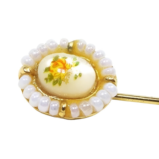 Front view of the top of the Mid Century vintage floral stickpin. The metal is gold tone in color. The top part is oval in shape and has a domed mother of pearl shell cab in the middle. There is a decal on top of the mother of pearl with a flower design in yellow and orange with green leaves. The outer edge of the oval has small pearly glass seed beads with small gold tone beads at each side. 