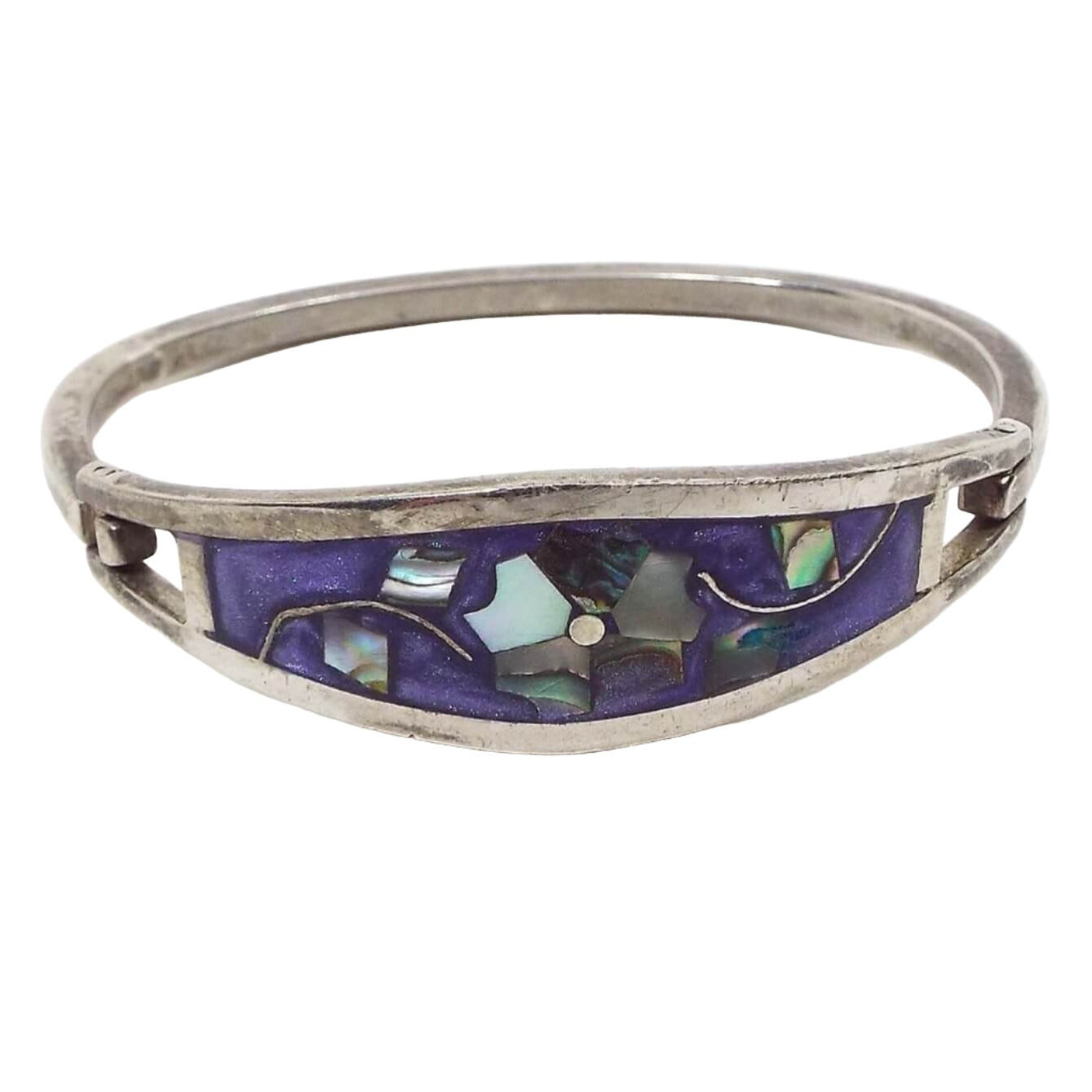 Angled front and top view of the retro vintage Taxco hinged floral bangle bracelet. The metal is silver tone in color. There is a large curved area on top with pearly purple enamel. In the middle is a flower design that has pearly multi colored inlaid abalone shell for the petals and leaves. The back of the bracelet is a squared bar that curves around and is hinged on one side and has a hook on the other to open and close the bracelet.