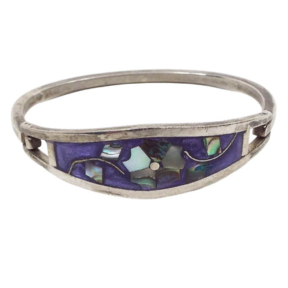 Angled front and top view of the retro vintage Taxco hinged floral bangle bracelet. The metal is silver tone in color. There is a large curved area on top with pearly purple enamel. In the middle is a flower design that has pearly multi colored inlaid abalone shell for the petals and leaves. The back of the bracelet is a squared bar that curves around and is hinged on one side and has a hook on the other to open and close the bracelet.