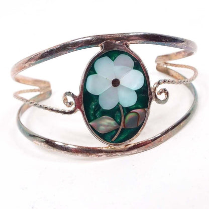 Front view of the Mexican Alpaca cuff bracelet has a large oval piece in the middle with pearly green resin with a flower design in the middle. Petals of the flower are pearly white mother of pearl and the leaves are pearly multi color abalone shell. there is a band of silver color metal above and below the oval that curve around to form the cuff. There is another thinner band of metal in the middle that comes off each side of the oval and is twisted and curled at the ends.
