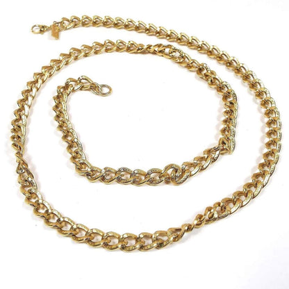 Top view of the retro vintage Monet chain necklace. The metal is gold tone in color. The chain has textured curved oval curb links with a dot pattern on them. There is a hinged clip clasp at the end that has a rectangle hang tag by it with Monet stamped on it.