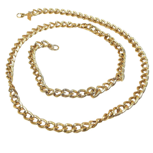 Top view of the retro vintage Monet chain necklace. The metal is gold tone in color. The chain has textured curved oval curb links with a dot pattern on them. There is a hinged clip clasp at the end that has a rectangle hang tag by it with Monet stamped on it.