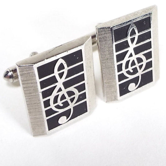Angled front and side view of the retro vintage Swank musical note cufflinks. The metal is silver tone in color. There are faceted rectangles on the front that have a treble clef design with a black painted background. Part of the rounded end levers on the back can be seen.
