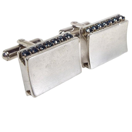 Angled front and side view of the Mid Century vintage rhinestone cufflinks. The metal is silver tone in color. The fronts have a slightly curved indented shape. The sides have a channel set row of dark blue rhinestones and the backs have rounded bar style levers.