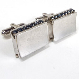 Angled front and side view of the Mid Century vintage rhinestone cufflinks. The metal is silver tone in color. The fronts have a slightly curved indented shape. The sides have a channel set row of dark blue rhinestones and the backs have rounded bar style levers.