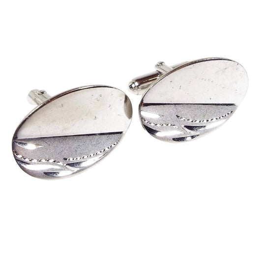 Angled front view of the Mid Century vintage Anson sterling silver cufflinks. They are brighter silver in color and oval in shape. There have two diagonal sections on the oval with one being plain shiny silver and the other being matte silver with a shiny e