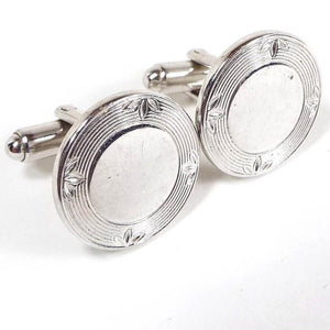 Angled front and side view of the Mid Century vintage cufflinks. The fronts are round with shiny silver tone metal middle areas. there is a line design around the edge that has sets of engraved leaves at each side. The rounded ends of the levers on the back can be seen in the photo.