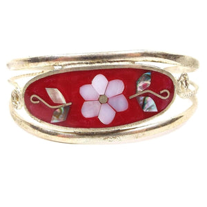 Front side of the Mexican Alpaca cuff bangle. It has a curved wide oval on the front with red resin enamel and a white pearly mother of pearl shell flower with multi color abalone shell leaves on each side. Metal is silver in color and has a band above and below the oval that curves to make the rest of the bracelet. There is a thinner twisted band in the middle that is curled at either side of the red oval.