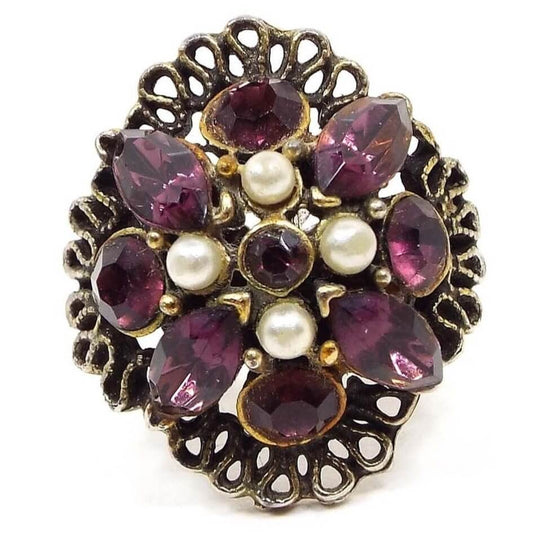 Top view of the Mid Century vintage rhinestone and faux pearl adjustable ring. The metal is antiqued brass in color. The top part has a wide filigree base with cut out teardrop style design. There is a small round bezel set dark purple rhinestone in the middle surrounded by four plastic faux pearls. The outer edge of the design has alternating bezel set round and marquis shaped dark purple rhinestones. 