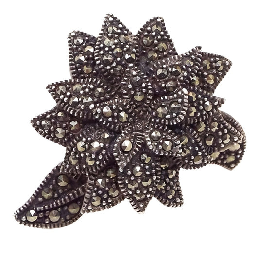 Top view of the large retro vintage sterling silver marcasite flower ring. The sterling has darkened with age for a dark gray color. The floral design has three layers of petals all the way around and three leaves curved off the sides. Each petal and leaf has tiny round metallic gray pavé set marcasite stones. 
