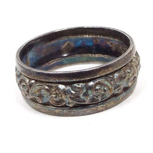 Angled top and side view of the Mid Century vintage Clark and Coombs band ring. The sterling silver is very darkened from age for a darkened gray color. There are some metallic blue areas seen on the inside of the band. The outside of the ring has a line around the top and bottom edge with a raised leaf design all the way around the middle of the band.
