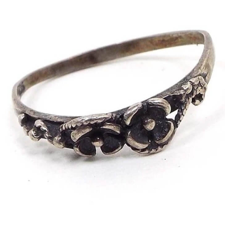 Angled front view of the retro vintage sterling silver floral ring. It is darkened silver in color with a thinner sized band. The top part of the ring has a small raised flower design. The band is slightly bent so it's not perfectly rounded on the back.