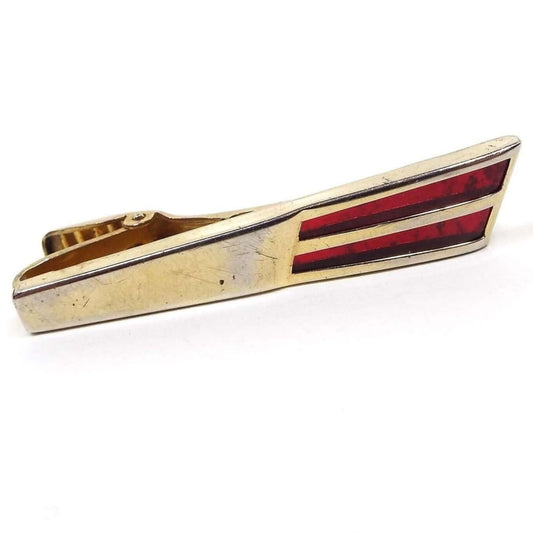 Front view of the Mid Century Anson vintage tie clip. It has a Modernist style flared gold tone metal bar with two flared stripes of inlaid lucite plastic at the end in a deep red color. There is some minor scruff scratching and an area in the middle that is slightly lighter on the metal from age. 