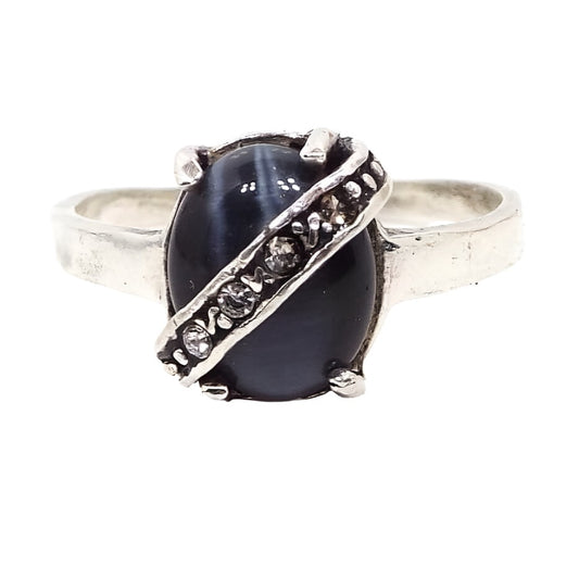 Top view of the retro vintage faux cat's eye ring. The glass imitation cat's eye cab is oval in shape and dark gray in color. It has ribbons of flashy lighter gray as you move around. There is a band of rhinestones running diagonally across the glass cab. The rhinestones are prong set inside a channel. The band and setting of the ring are silver tone in color.