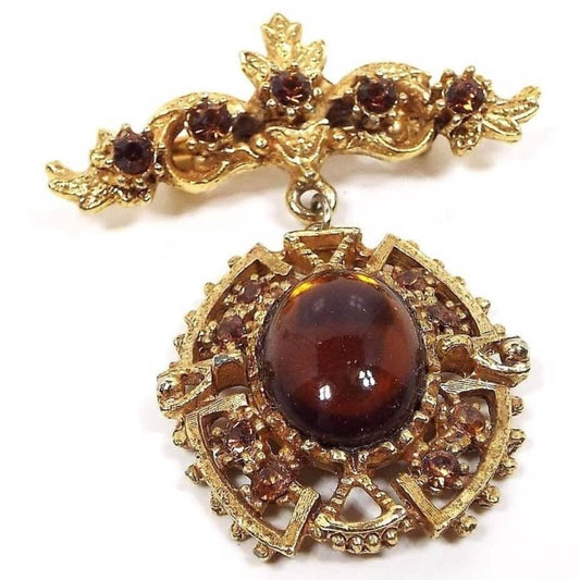 Front view of the 1960's Mid Century vintage Victorian revival dangle brooch pin. The metal is textured matte gold tone in color. The top bar has 5 small prong set burnt orange rhinestones surrounded by curls, small leaves, and a small flower in top. The bottom drop dangle is larger and has an oval bezel set burnt orange glass cab. There are filigree curved elbow and triangle shapes around the cab. The curved shapes each have two more small round prong set burnt orange rhinestones. 