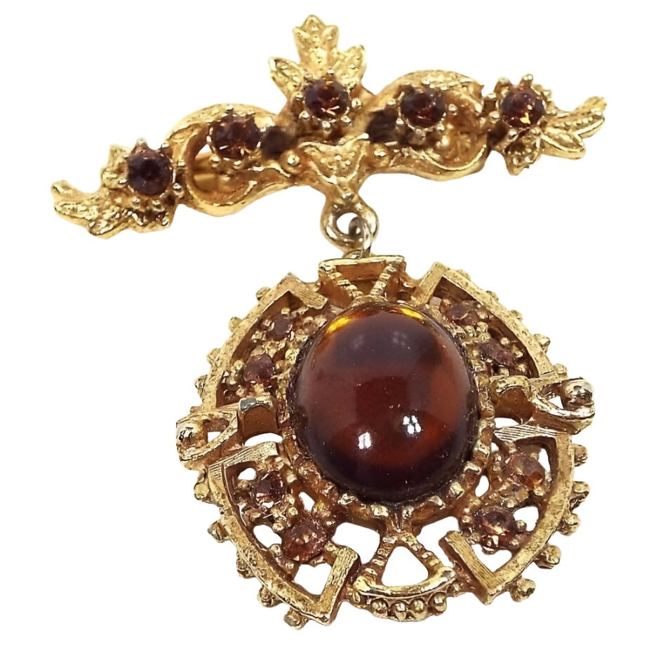 Front view of the 1960's Mid Century vintage Victorian revival dangle brooch pin. The metal is textured matte gold tone in color. The top bar has 5 small prong set burnt orange rhinestones surrounded by curls, small leaves, and a small flower in top. The bottom drop dangle is larger and has an oval bezel set burnt orange glass cab. There are filigree curved elbow and triangle shapes around the cab. The curved shapes each have two more small round prong set burnt orange rhinestones. 